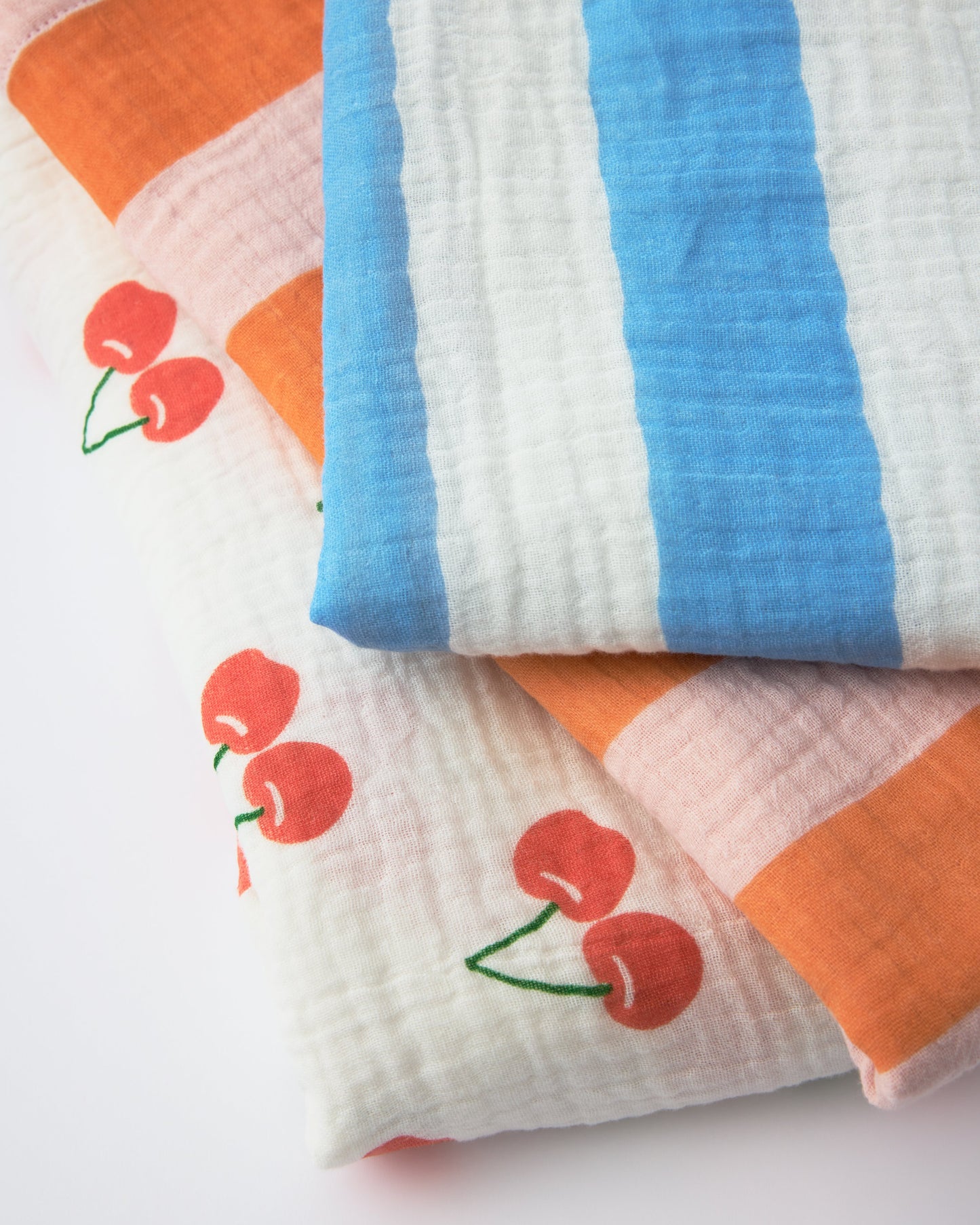 Three patterns of Organic Muslin Burp Cloth (Three-Pack) stacked, Blue Wide Stripe, Pink Wide Stripe, White Cherries, on a beige background.