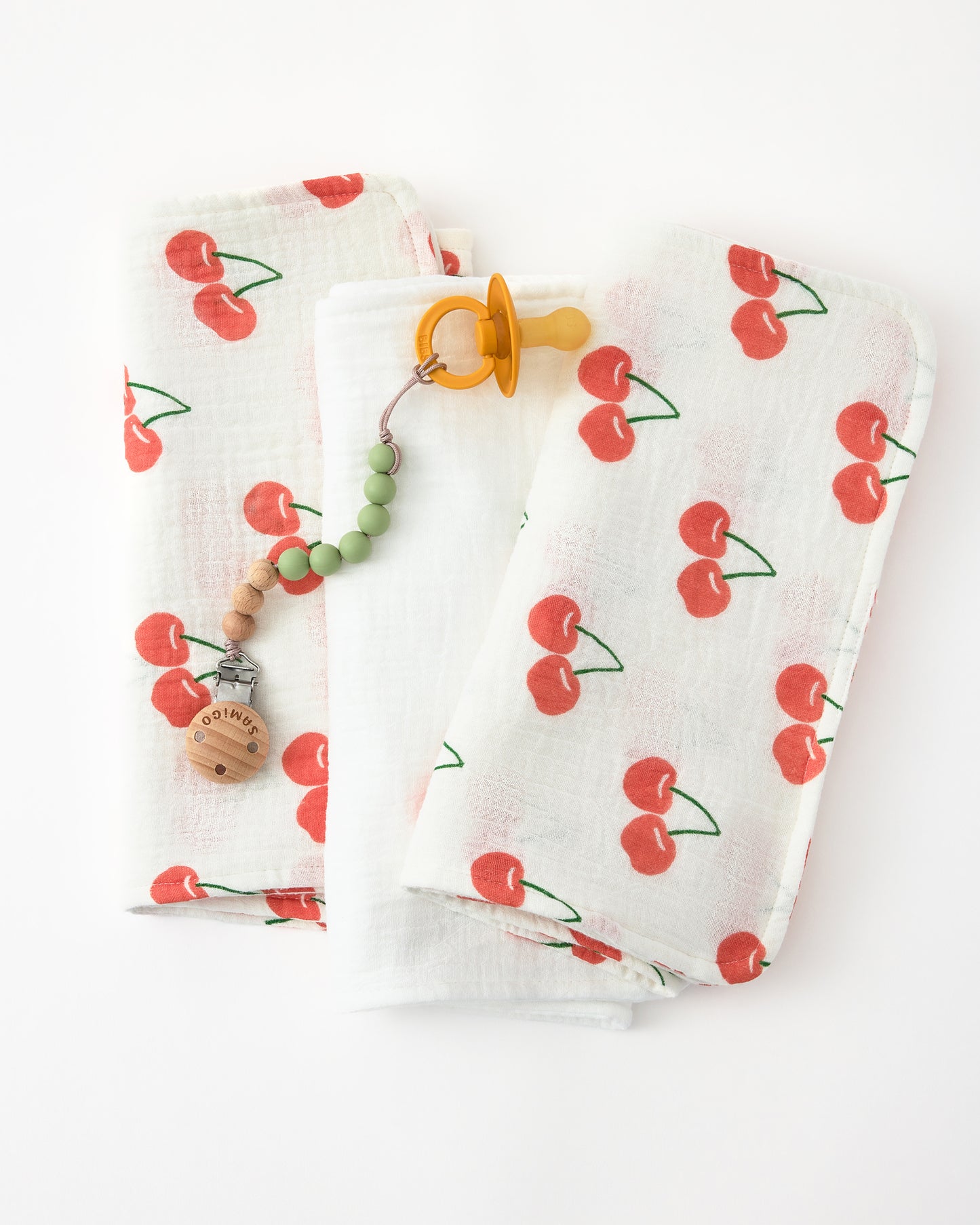 A baby pacifier on a chewtoy tether resting on Organic Muslin Burp Cloth (3-Pack) in White Cherries on a white background.