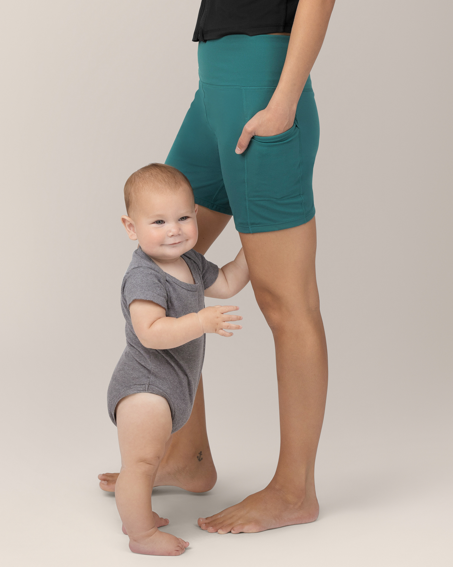 Side view of model wearing the Stevie Active Postpartum Bike Short in Teal while a baby stands beside her