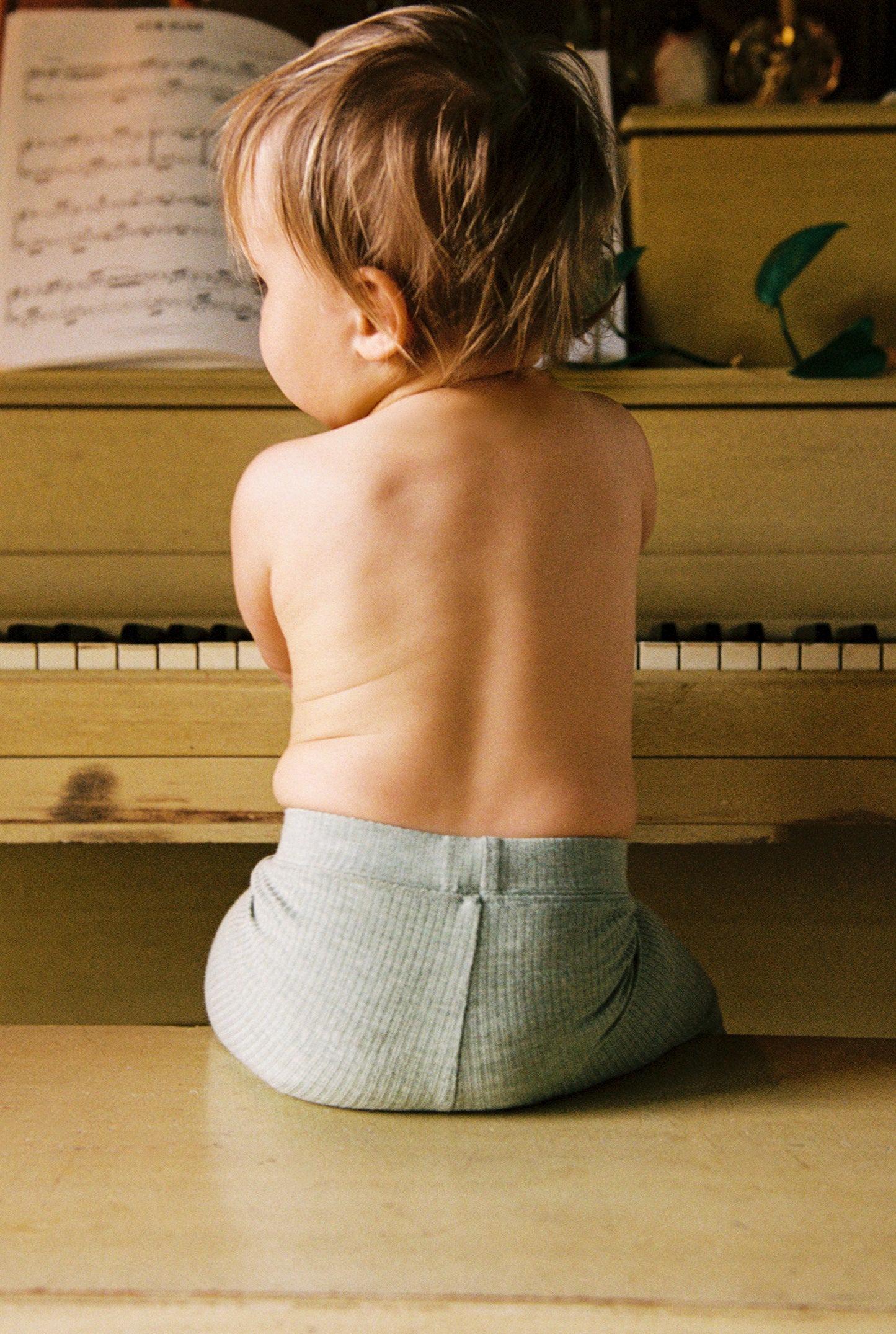 Baby wearing the bottoms from Ribbed Bamboo Baby Two-Piece Pajama Set in Sage Heather, sitting at piano