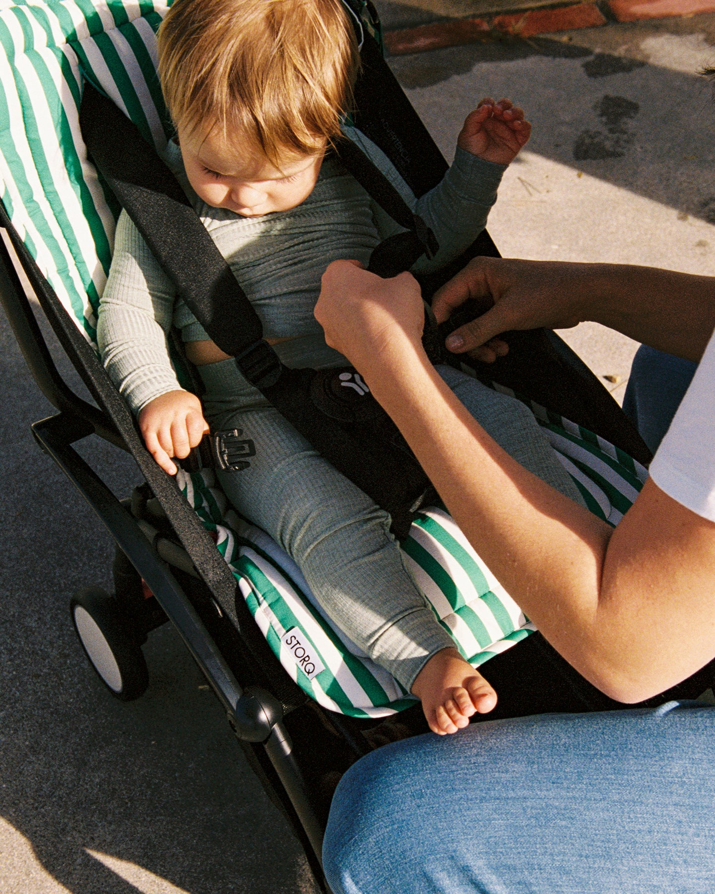 Child being secured in a stroller with a Stroller Party Seat Liner in Green Stripe, being fastened by an adult.