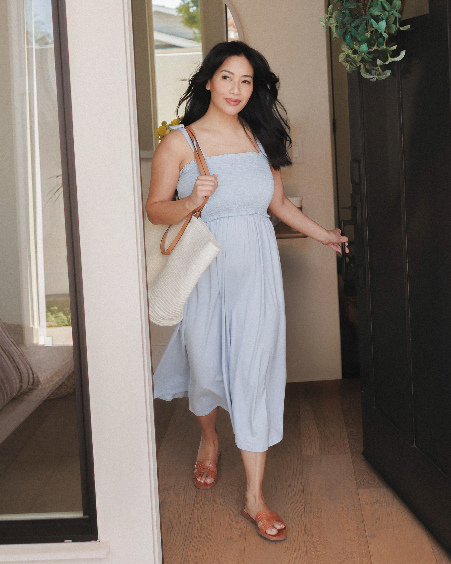 Model wearing the Sienna Smocked Maternity & Nursing Dress in French Blue, holding a purse, walking out of her house. @model_info:Nikki is wearing a Medium.