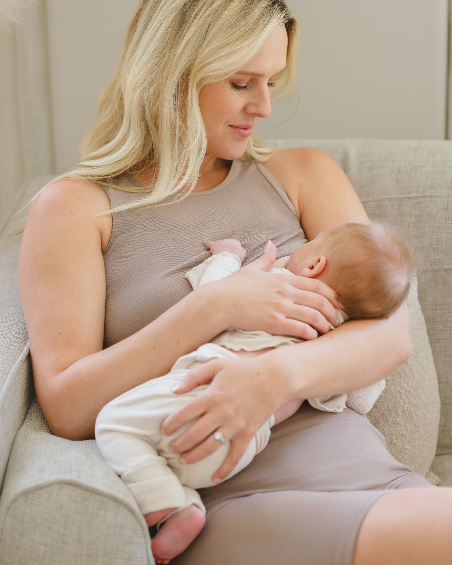 Model sitting on sofa cradling and nursing baby while wearing the Sublime® Bamboo Maternity & Nursing Longline Bra Top in Taupe, paired with matching Sublime® Bamboo Maternity & Postpartum Bike Short