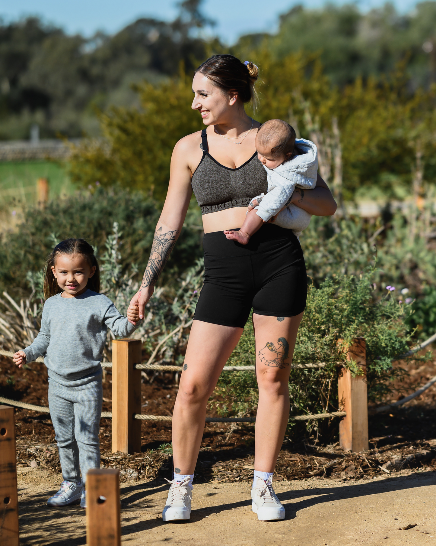 Model holding a baby outside while wearing the Stevie Active Postpartum Bike Short in Black with the Sublime Hands Free Pumping and Nursing Sports Bra @model_info:Anna is wearing a Small.