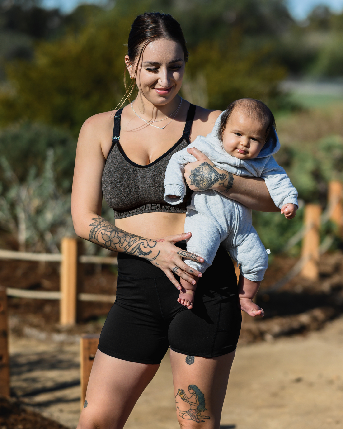 Model holding a baby outside while wearing the Stevie Active Postpartum Bike Short in Black with the Sublime Hands Free Pumping and Nursing Sports Bra
