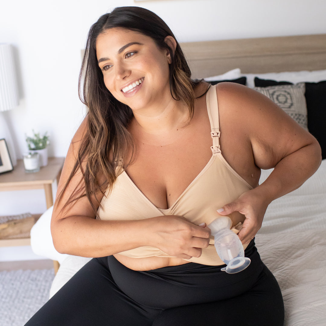 Model sitting on a bed wearing the Sublime® Hands-Free Pumping & Nursing Bra - Super Busty in Beige with a pump attached.