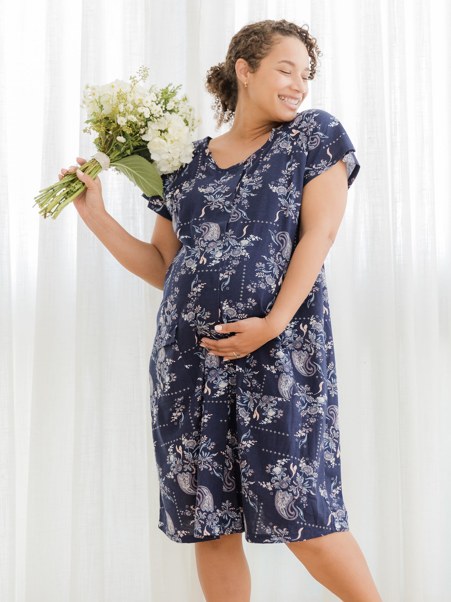 Front view of pregnant model wearing the Universal Labor & Delivery Gown in Navy Paisley, holding flowers in one hand @model_info:Alysha is 5'6" and wearing a S/M/L.