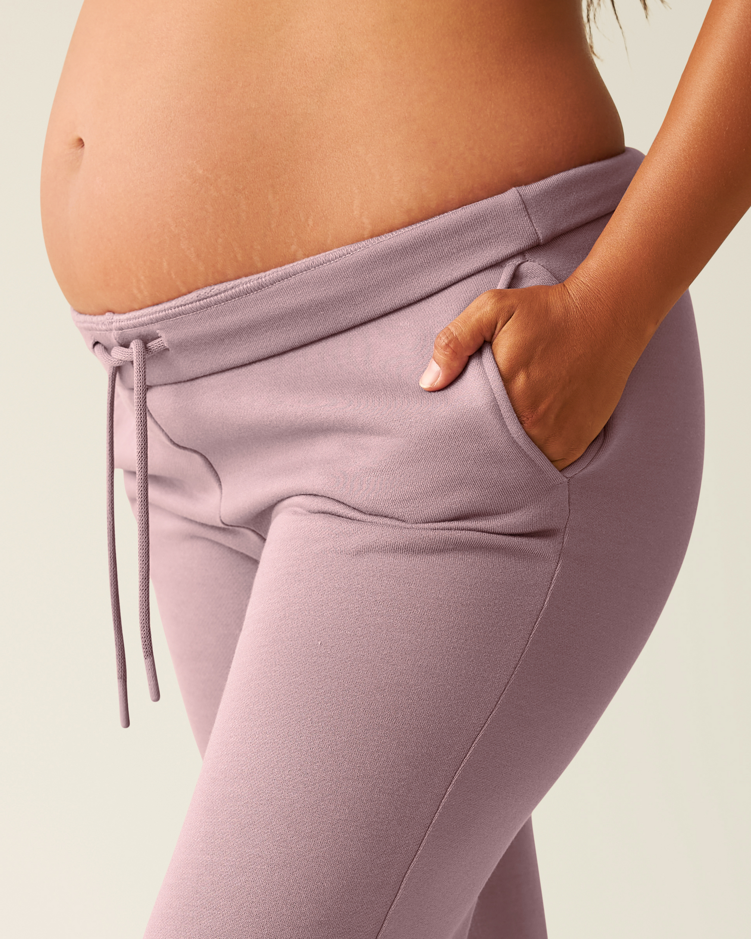 Close-up of woman wearing dusty mauve Cloud Fleece Sweatpant on a beige background, focusing on folded waistband and hand in pocket