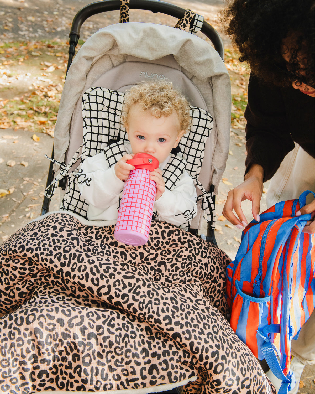 In a park setting, a baby is sitting in a beige stroller, on the Stroller Party™ Seat Liner & Strap Covers in Black Grid and snuggled under the Cozy Stroller Blanket in Leopard, while drinking from the Cool Kid Water Bottle in Orchid Grid. Mother is holding the Cool Kid Backpack in Blue & Rust Stripe.