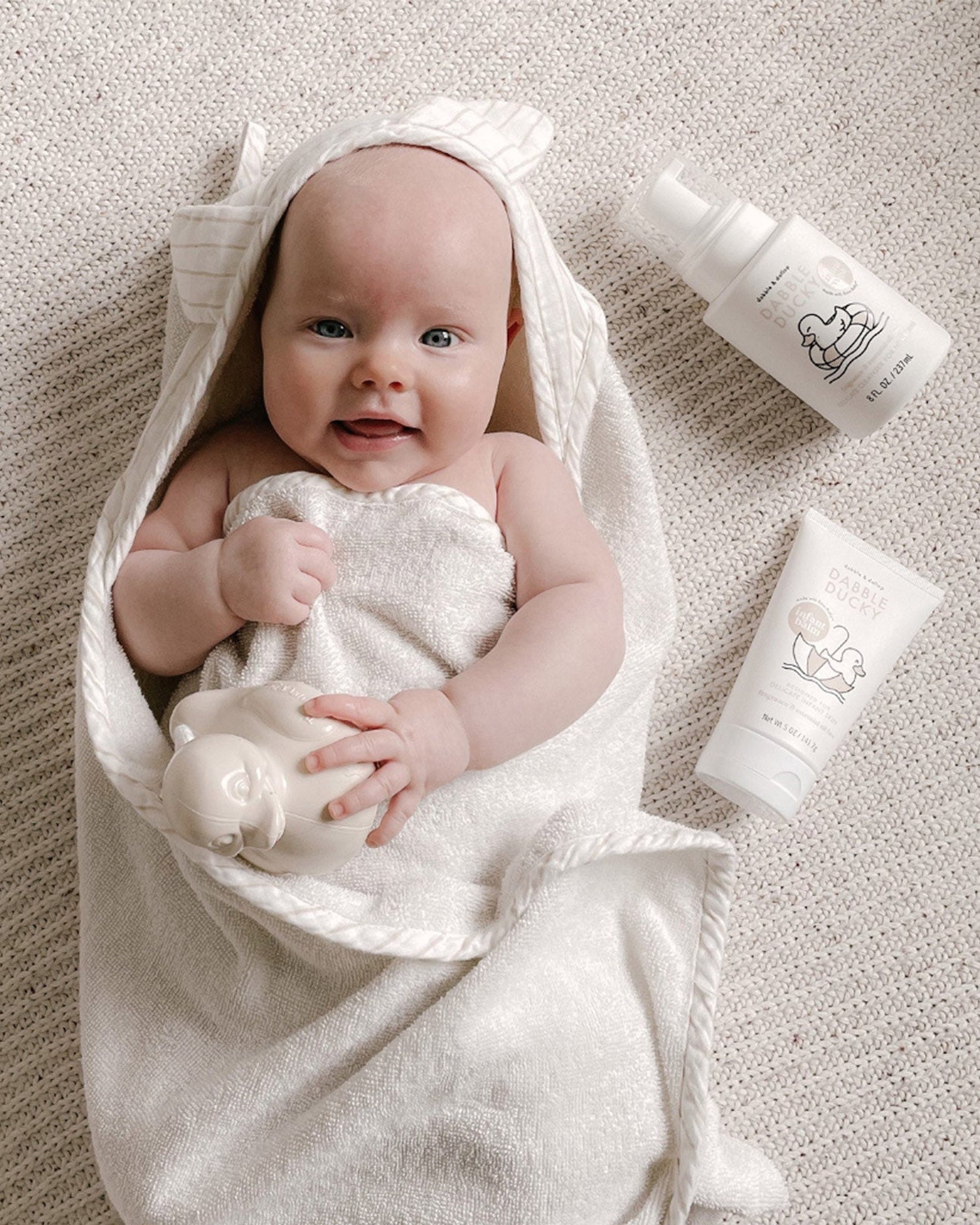 Baby wrapped in a white towel with Dabble Dollop Dabble Ducky infant wash and infant balm on beige background