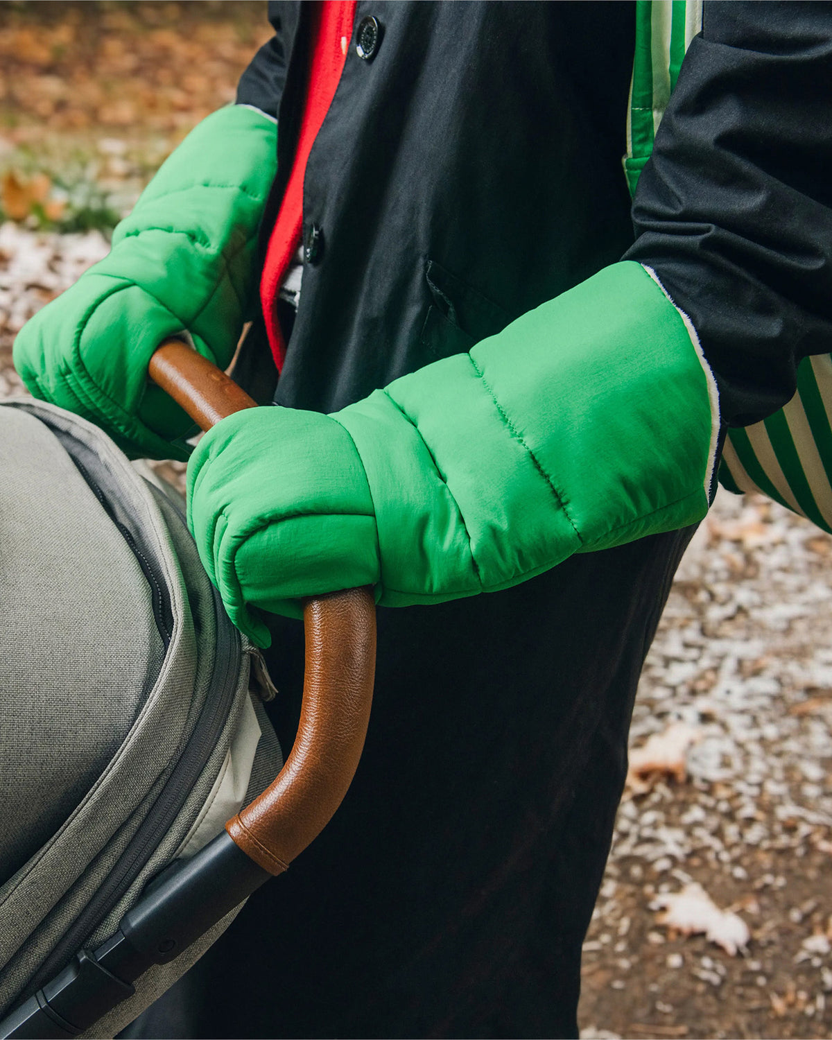Model is pushing grey stroller while wearing Cozy Stroller Mitts in Green, back coat in a natural setting with spots of snow on the ground.