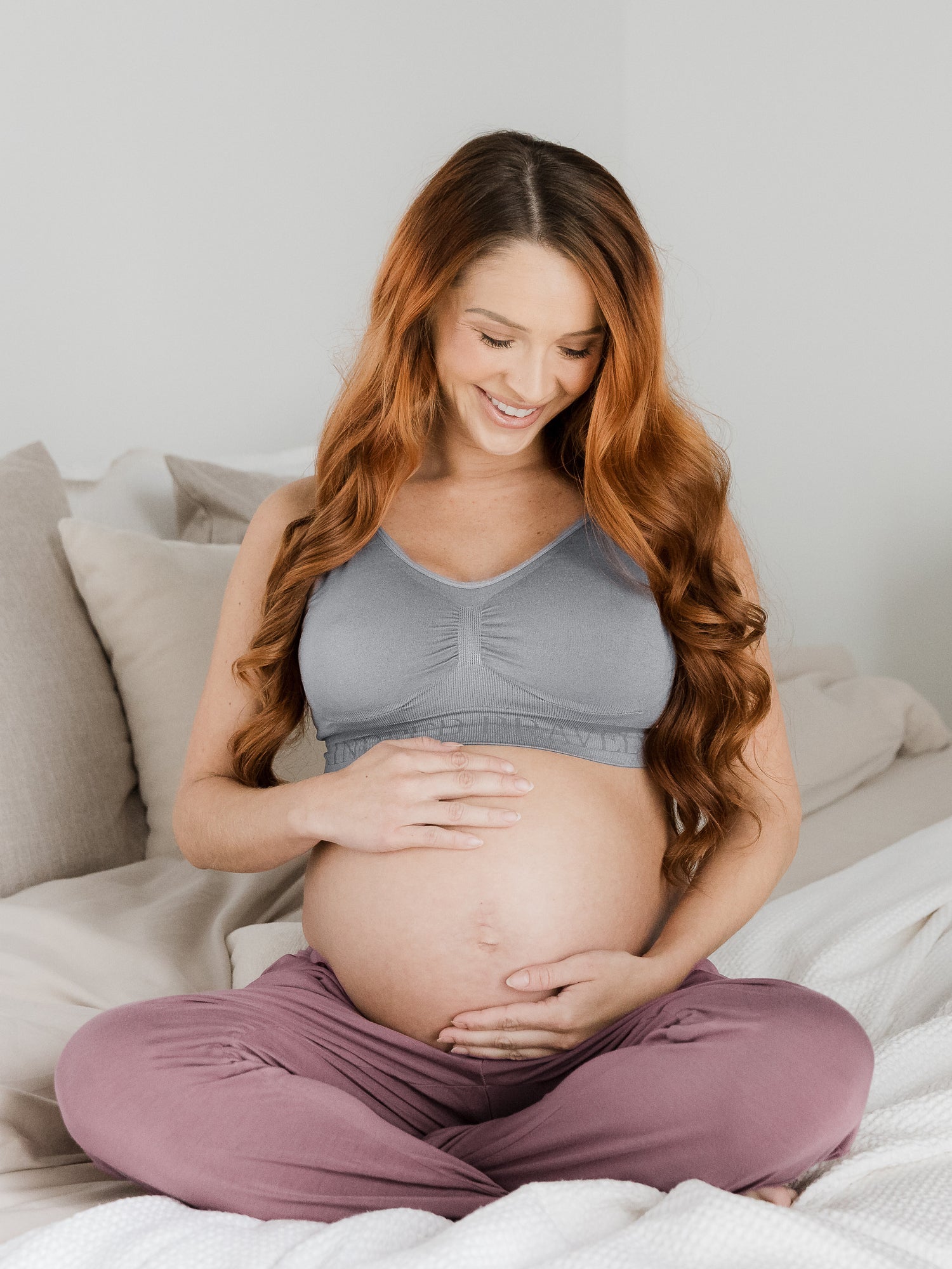 Pregnant model wearing the Simply Sublime® Nursing Bra | Grey sitting on a bed and looking down at her stomach. @model_info:Shannon is wearing a Small Busty.