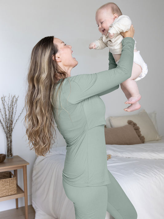 Model holding her baby up in the air while wearing the Jane Nursing Pajama Set in Sage. @model_info:Ali is wearing a Medium.