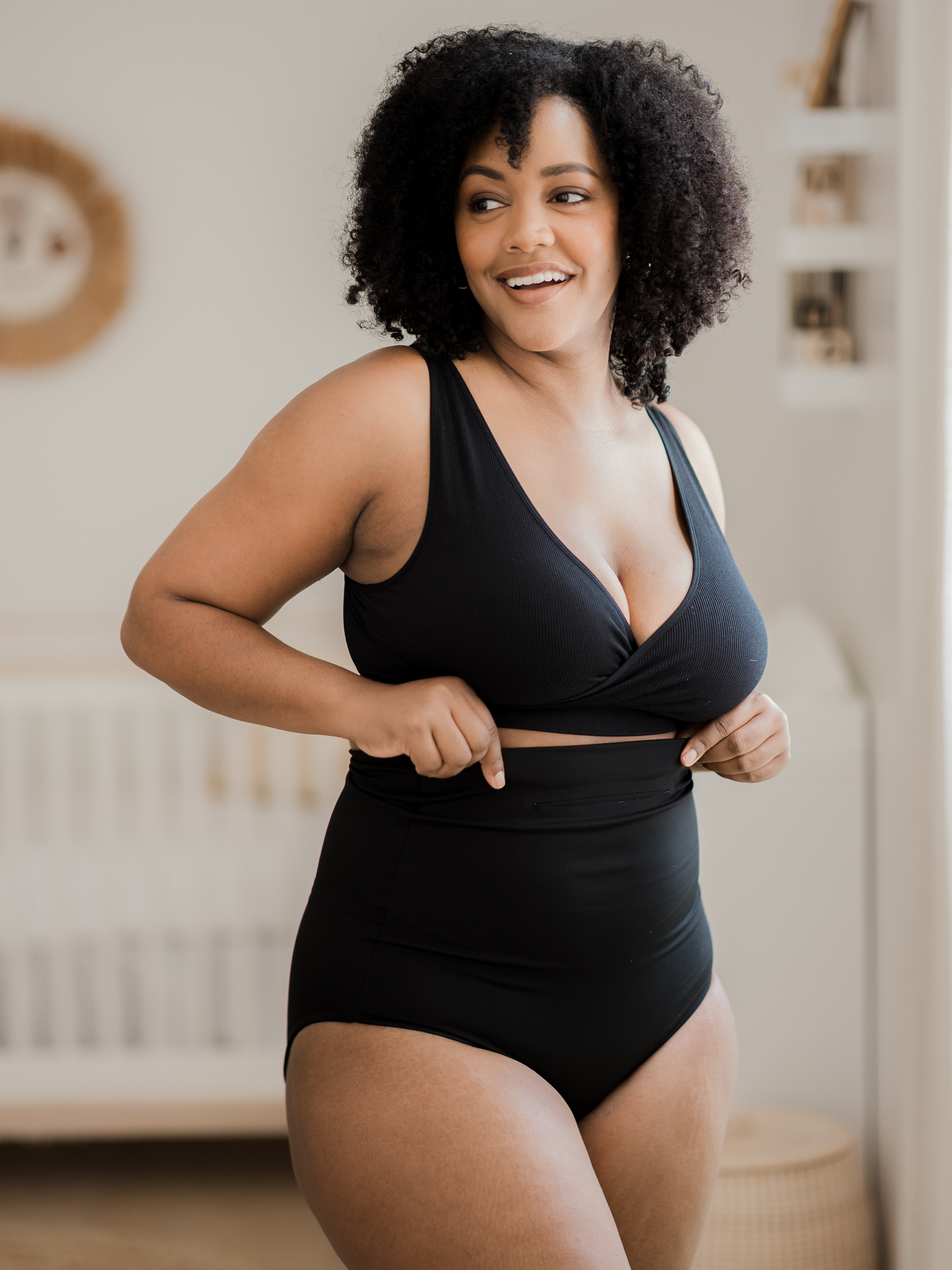 Model wearing the Soothing Fourth Trimester Underwear in Black standing in front of a crib. @model_info:Roxanne is 2 months postpartum and wearing a Large.