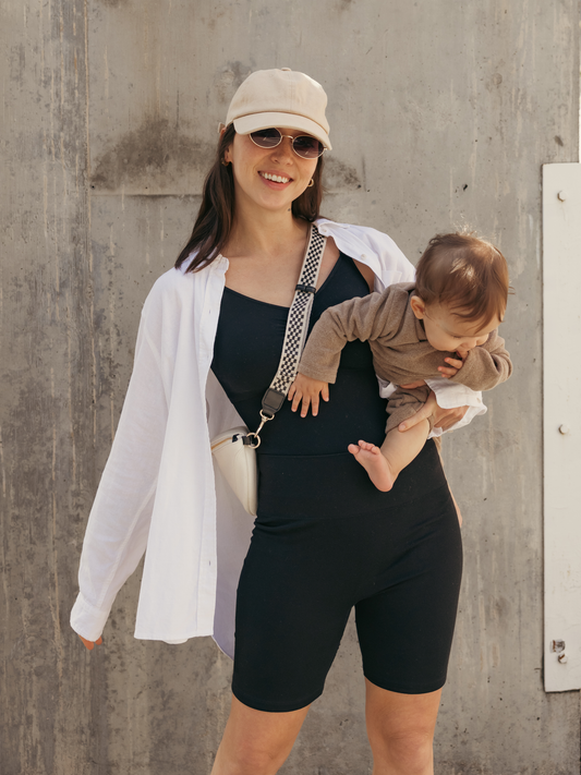 Model outside holding baby and wearing the Sublime® Bamboo Maternity & Postpartum Bike Short in black, paired with the Sublime® Bamboo Maternity & Postpartum Bodysuit and an open button-down shirt. @model_info:Lyn is 5'6" and wearing a Small.