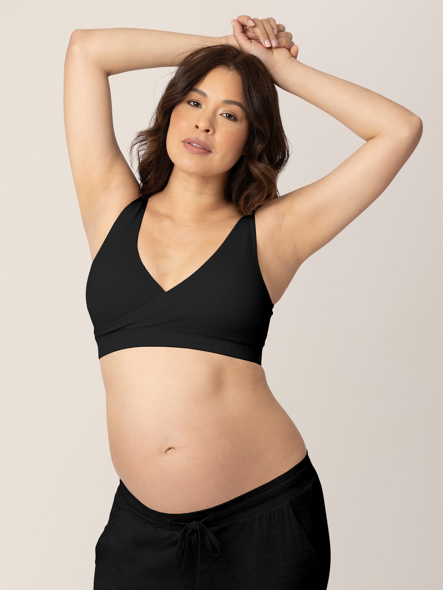 Pregnant model wearing the Sublime® Adjustable Crossover Nursing & Lounge Bra in Black with her hands above her head. @model_info:Vanessa is wearing a Medium.