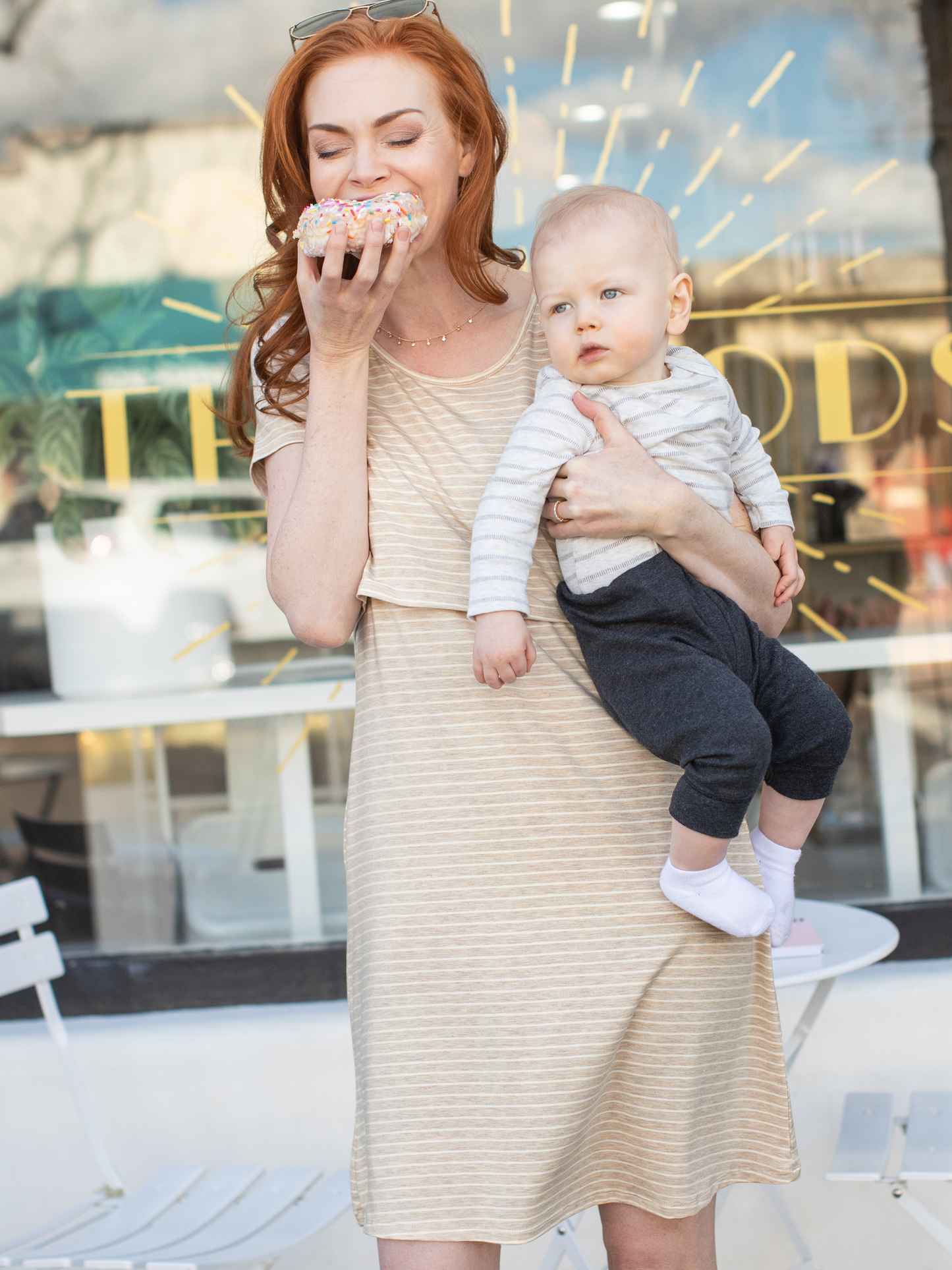 Model wearing the Eleanora Bamboo Maternity & Nursing Dress in Oatmeal Stripe biting into a donut while holding her baby on her hip. Her baby is looking wistfully up at the donut. @model_info:Sarah is wearing a Small.