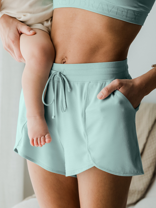 3/4 angle view of model wearing the Bamboo Maternity & Postpartum Lounge Shorts in dusty blue green, holding baby@model_info:Kate is wearing a Small.