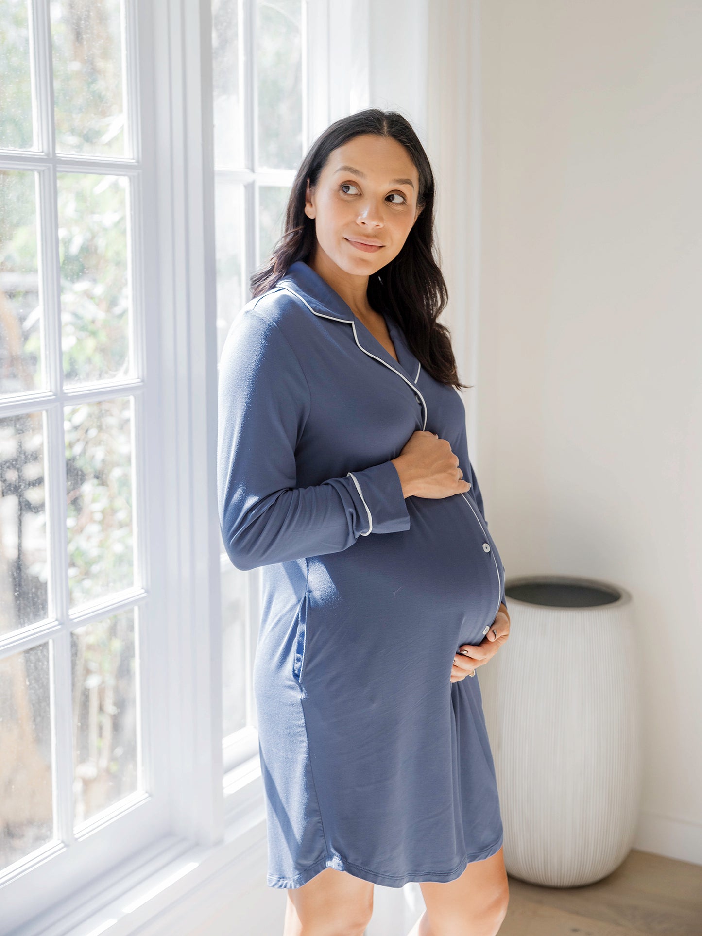 Model wearing the Clea Bamboo Long Sleeve Sleep Shirt in Slate Blue holding her pregnant belly standing in front of a window. @model_info:Julana is 5'9" and wearing a Small.