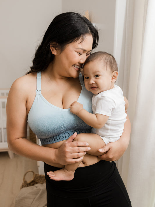 Model wearing the Sublime® Nursing Sports Bra in Ombre Storm holding her baby on her hip.@model_info:Binc is wearing a Medium Busty.