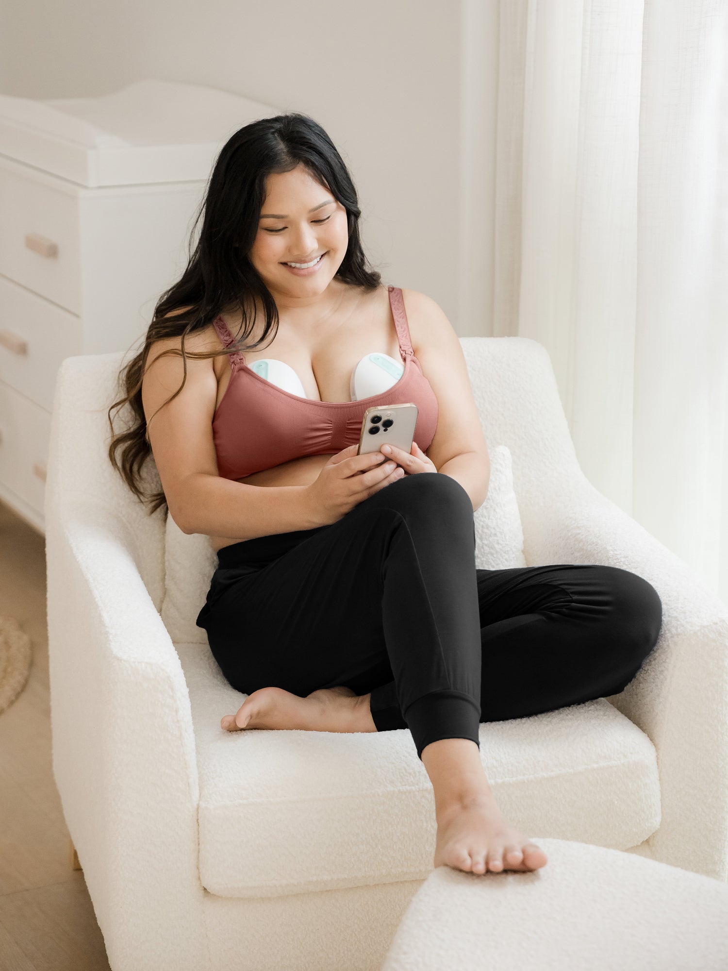 Model sitting on a chair while wearing the Simply Sublime® Nursing Bra in Redwood on her phone. @model_info:Binc is wearing a Medium Busty.
