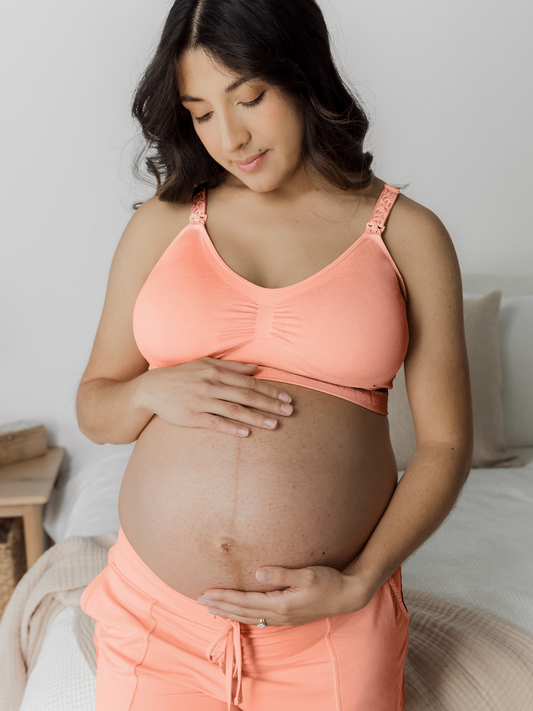 Pregnant model wearing the Simply Sublime® Nursing Bra in Vintage Coral holding her pregnant belly. @model_info: Patty is wearing a Medium Busty.