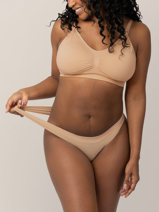 Model wearing the Grow with Me™ Maternity & Postpartum Thong in Beige holding onto the stretchy waistband on the thong. @model_info:Rashé is wearing a Medium. 