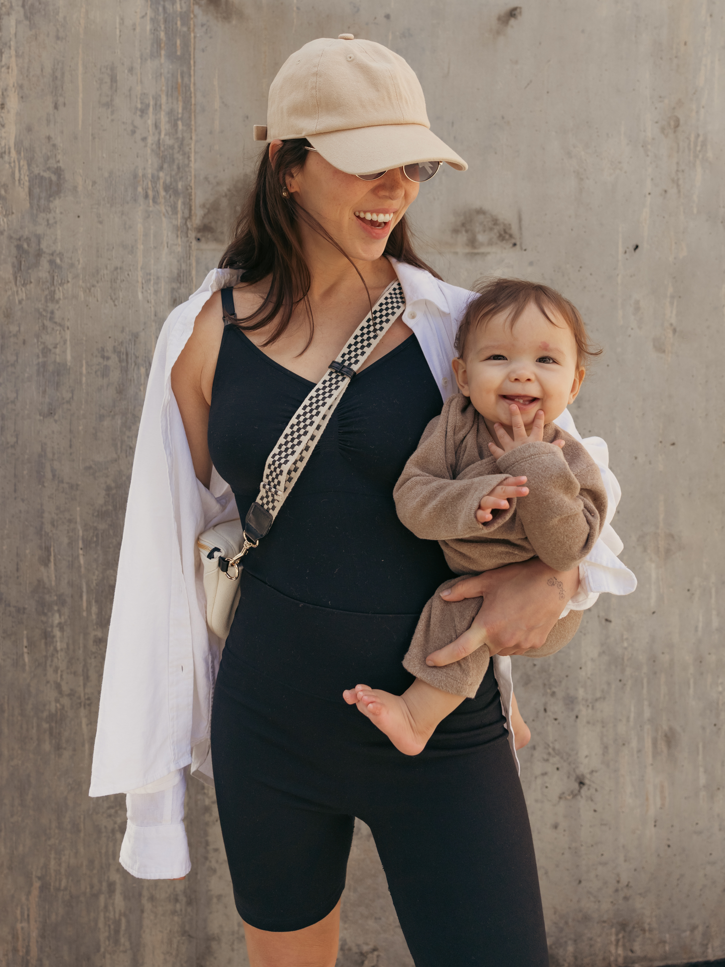 Model outside holding baby and wearing the Sublime®️ Bamboo Maternity & Nursing Bodysuit, paired with the Sublime® Bamboo Maternity & Postpartum Bike Short in black and an open button-down shirt..