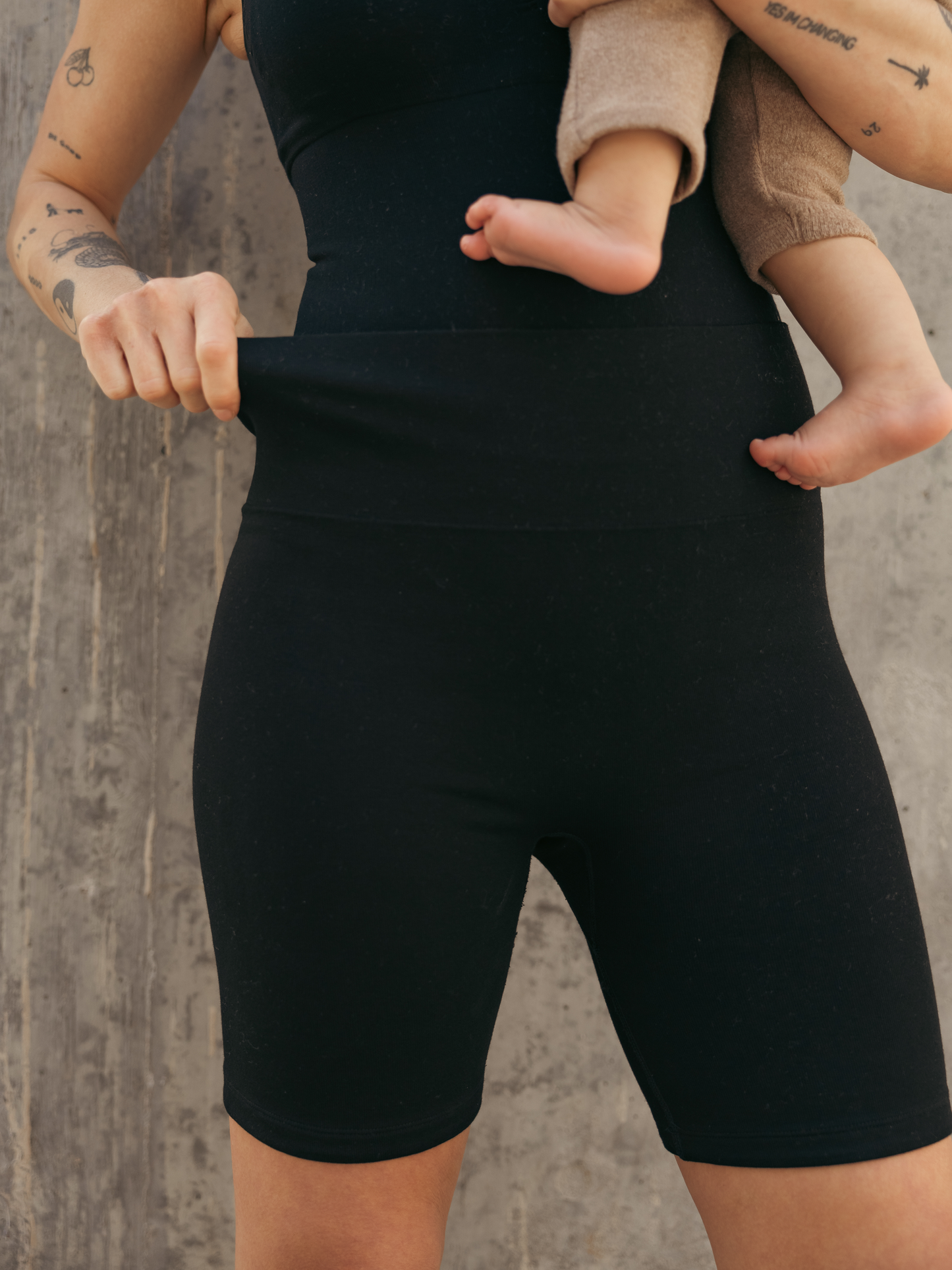 Close up of the Sublime® Bamboo Maternity & Postpartum Bike Short in black on model outside holding baby, showing stretch of short material.