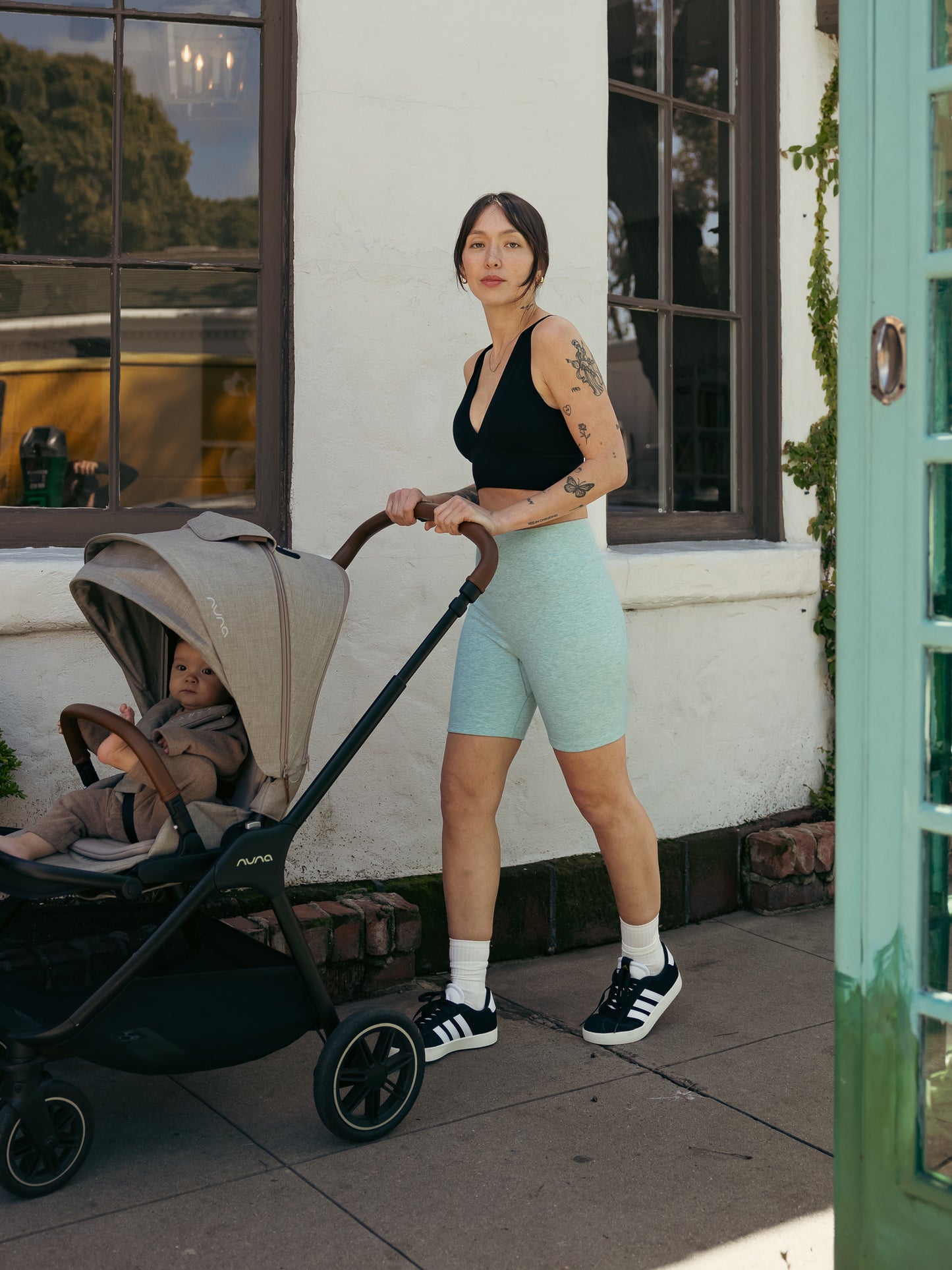 Model wearing the Sublime Maternity & Nursing Plunge Bra in Black paired with the Sublime Bamboo Bike Shorts in Dusty Blue Green. She is pushing her baby in a stroller.