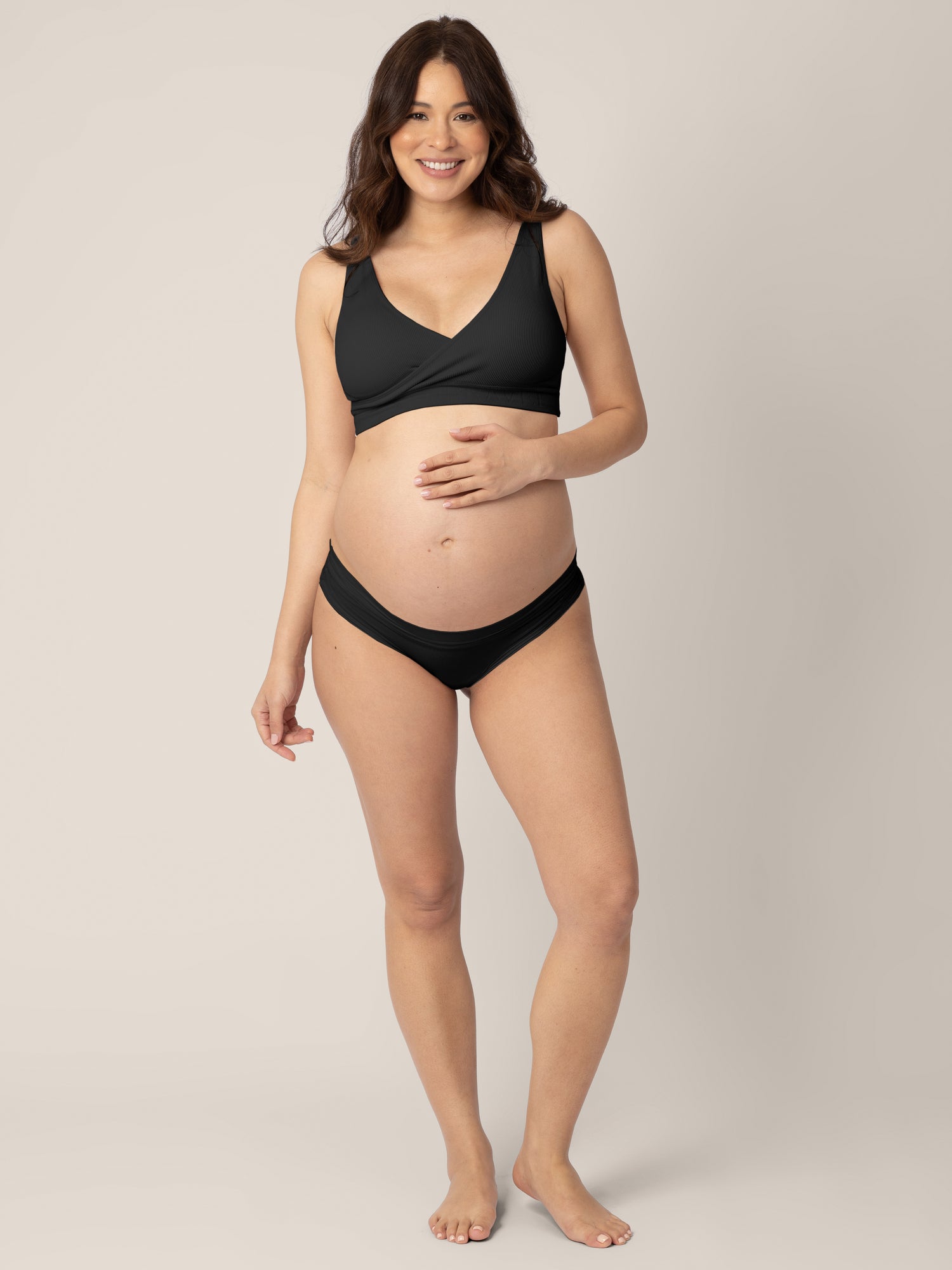 Pregnant model wearing the Grow with Me™ Maternity & Postpartum Thong in Black holding her belly bump. 