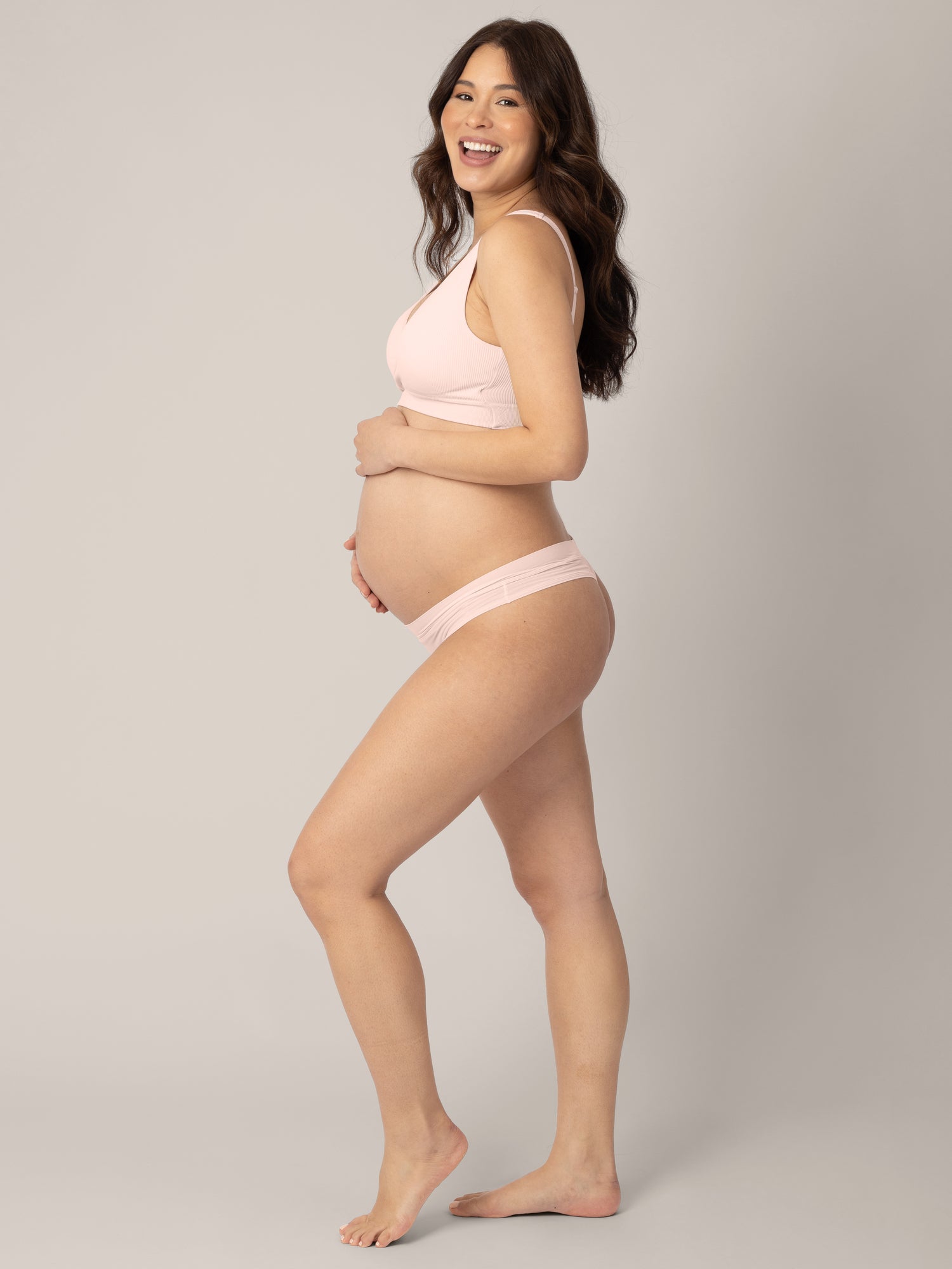 Pregnant model with her hand on her stomach and her other hand at her side smiling at the camera and wearing the Grow with Me™ Maternity & Postpartum Thong in Soft Pink.