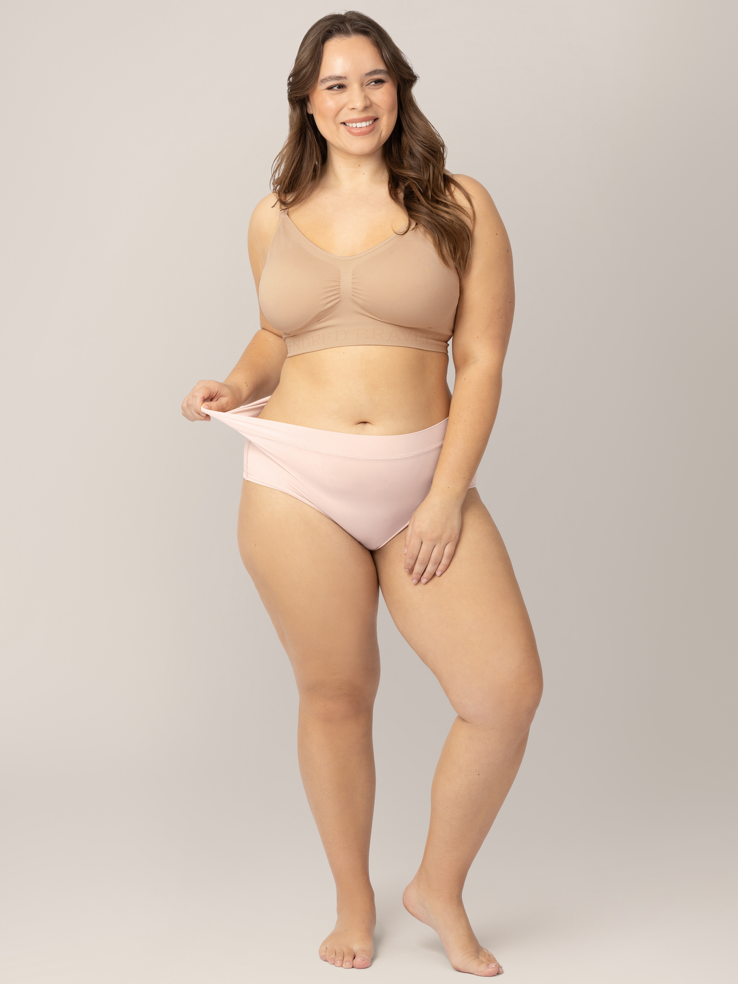 Model wearing the Grow with Me™ Maternity & Postpartum Brief in Soft Pink holding onto the waistband to show the elasticity. 