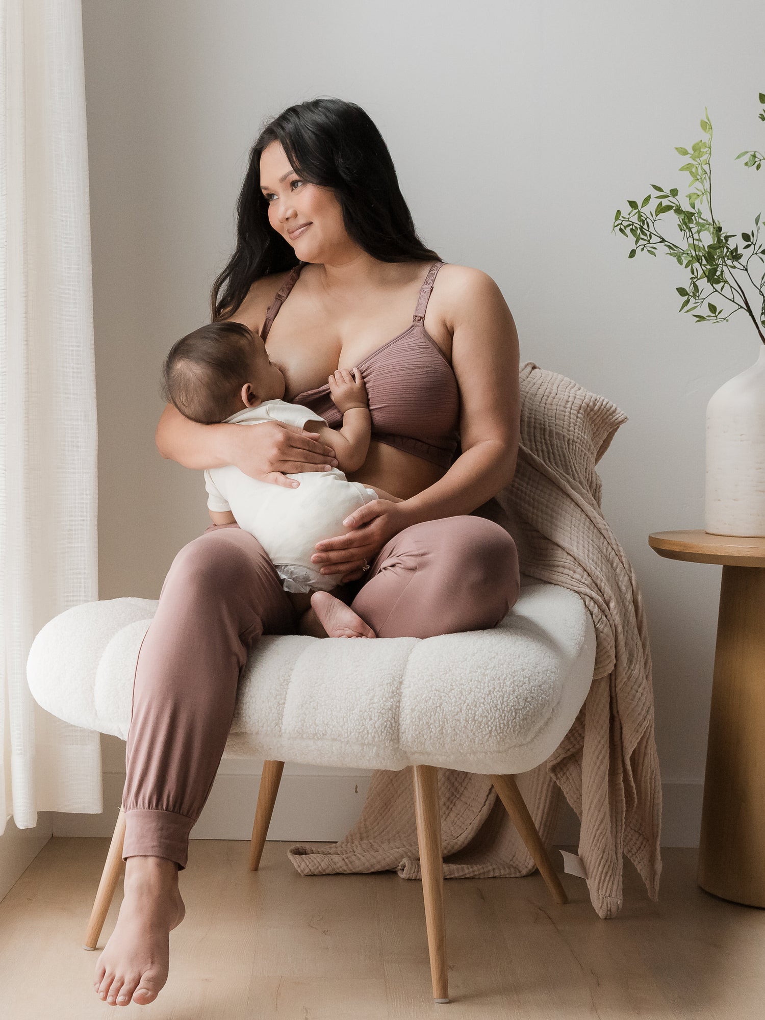 Model breastfeeding her baby while sitting in a chair and wearing the Sublime® Hands-Free Pumping & Nursing Bra in Twilight