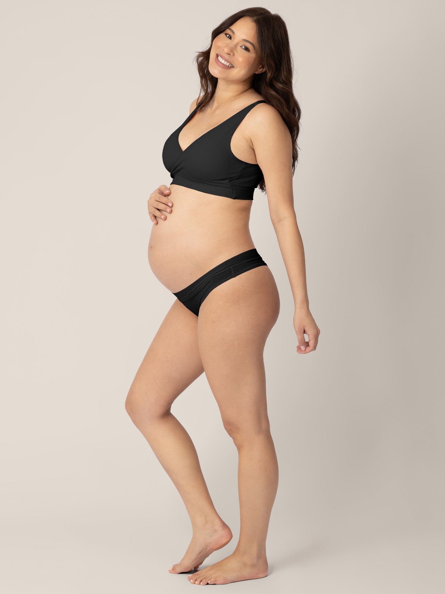 Pregnant model with her hand on her stomach and her other hand at her side smiling at the camera and wearing the Grow with Me™ Maternity & Postpartum Thong in Black