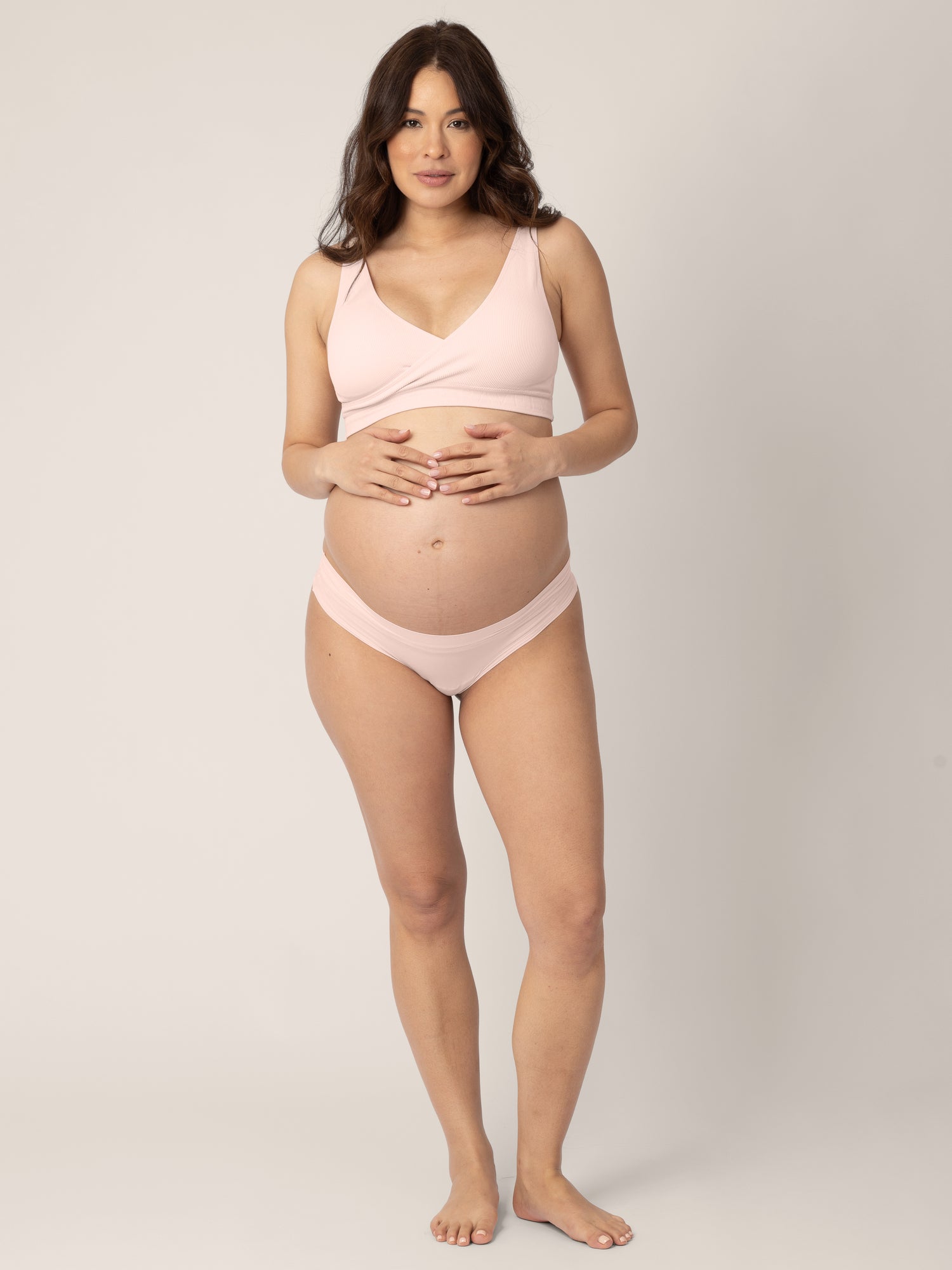 Pregnant model wearing the Grow with Me™ Maternity & Postpartum Thong in Soft Pink holding her belly bump.
