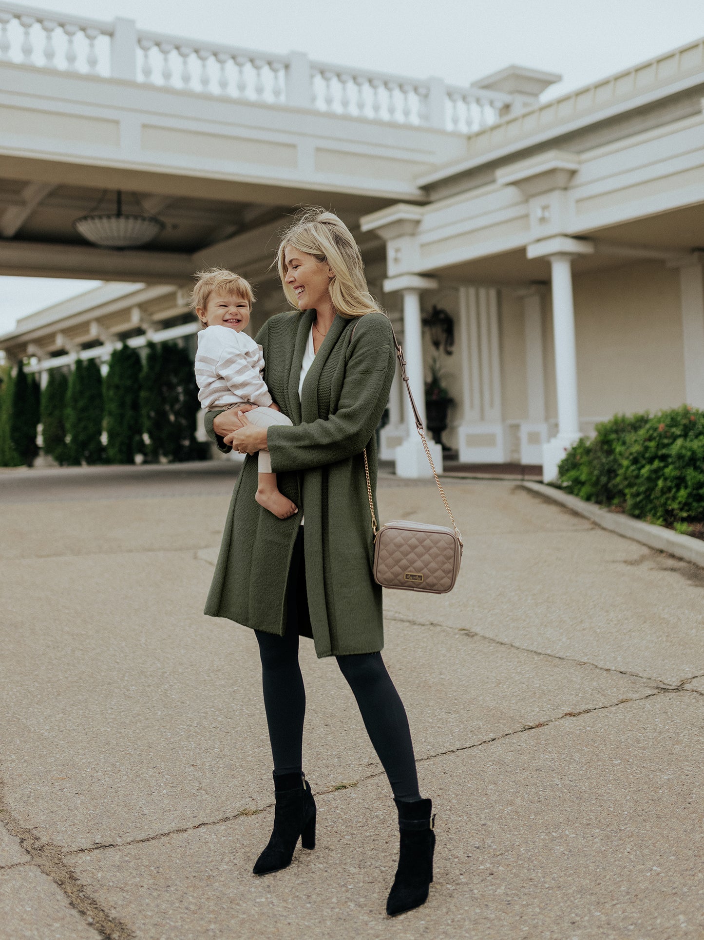 Model wearing the Chloe Cardigan Sweater in Thyme holding her baby standing in front of a fancy building. 