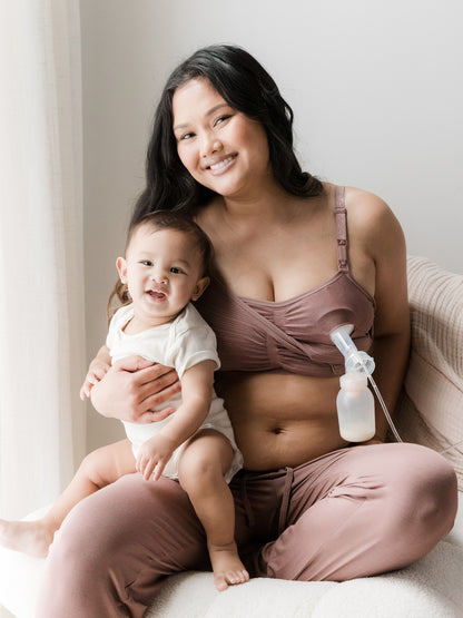 Model pumping while holding her baby on her lap and smiling wearing the Sublime® Hands-Free Pumping & Nursing Bra in Twilight