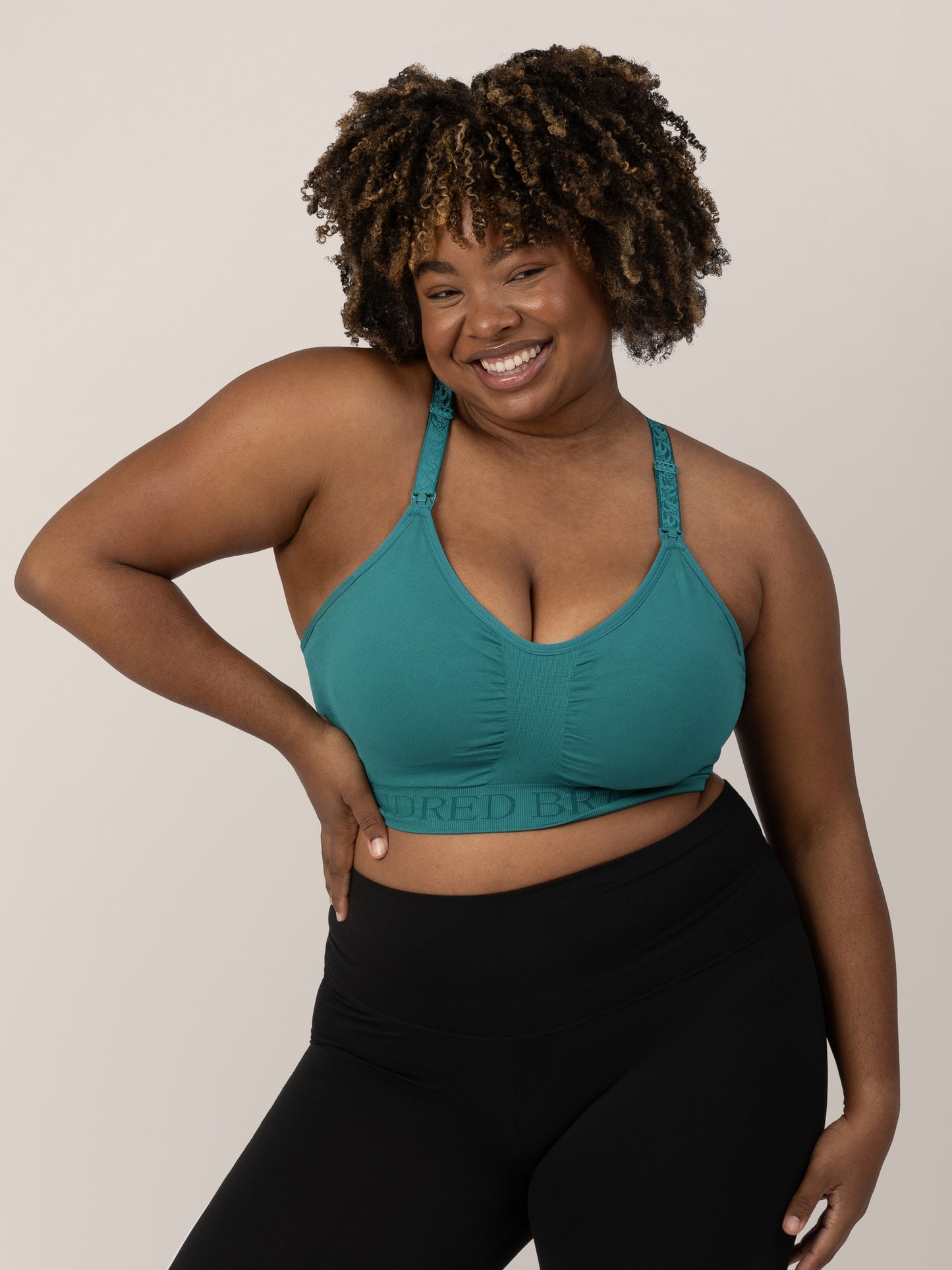 Model with her hand on her hip wearing the Sublime® Hands-Free Pumping & Nursing Sports Bra in Teal and smiling. @model_info:Skye is wearing an X-Large Busty.