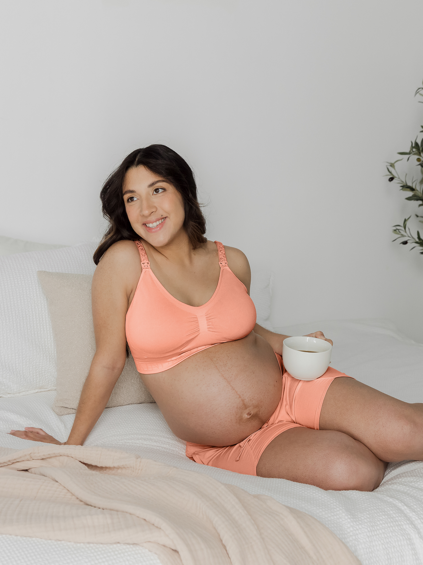 Pregnant model laying on a bed wearing the Simply Sublime® Nursing Bra in Vintage Coral with a cup of coffee.