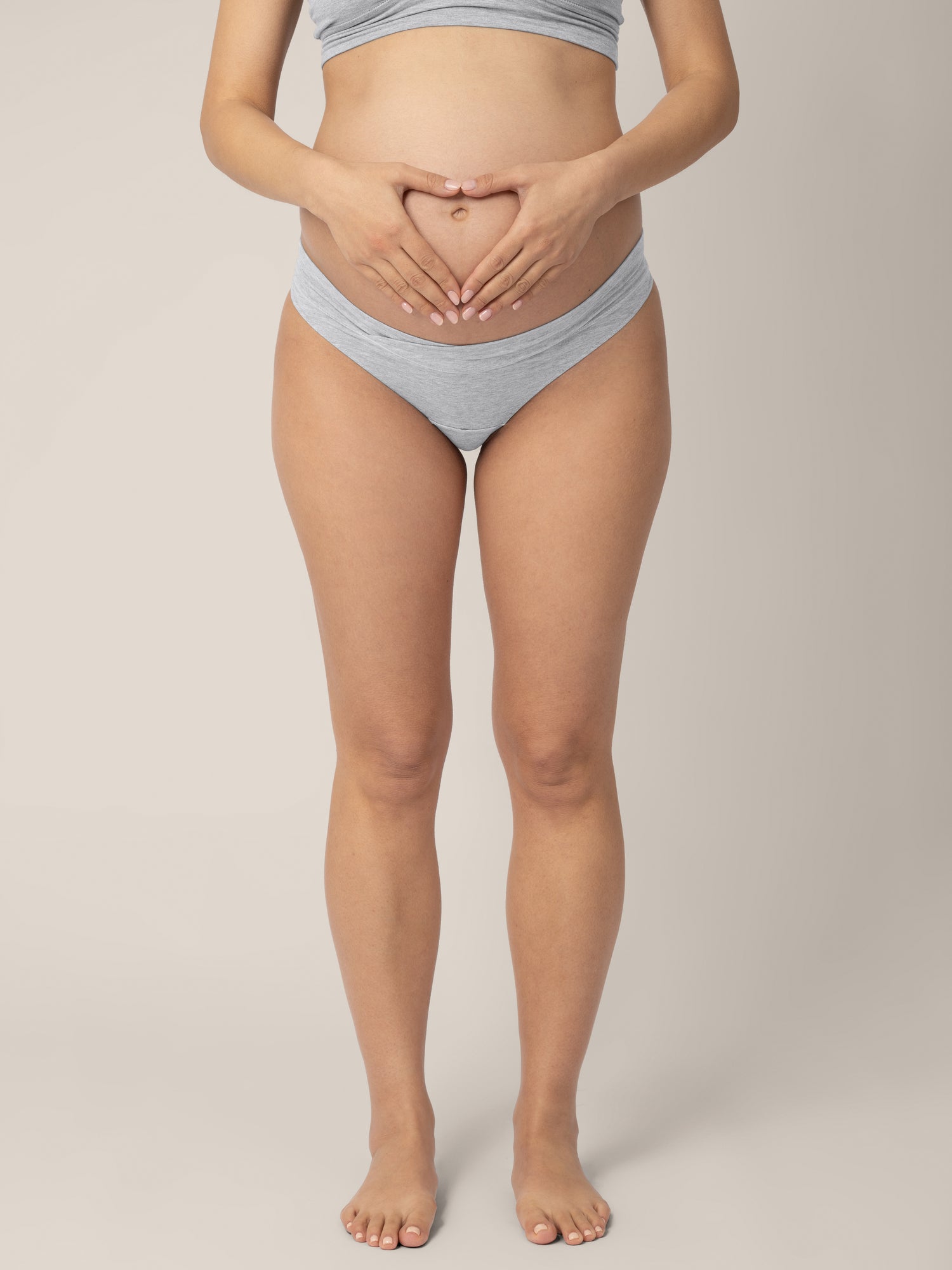 Bottom half of a pregnant model wearing the Bamboo Maternity & Postpartum Thong in Grey Heather making a heart on her belly.