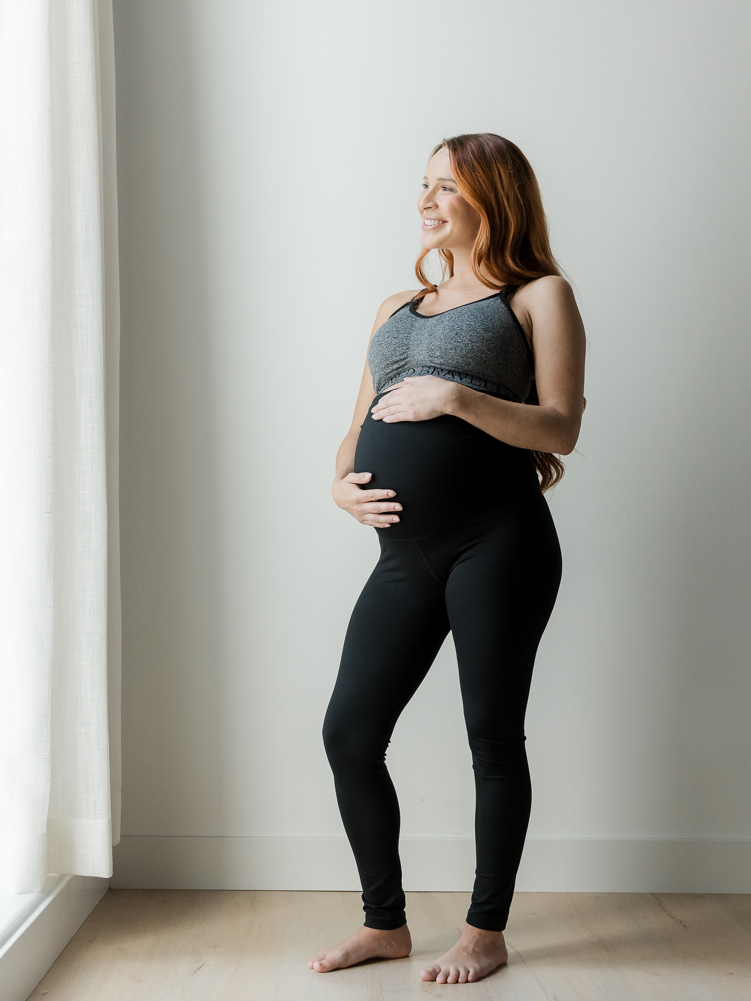 Pregnant model standing near a window holding her pregnant belly and wearing the Louisa Maternity & Postpartum Legging in Black. @model_info:Shannon is 5'8" and wearing a Small.
