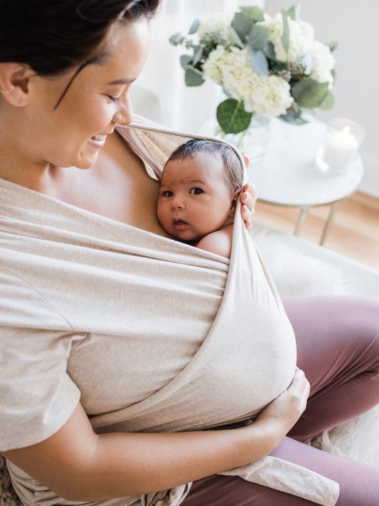 Model wearing the Organic Cotton Skin to Skin Wrap Top in Oatmeal Heather with her baby against her chest sitting in a chair.@model_info:Bri is wearing a Large.