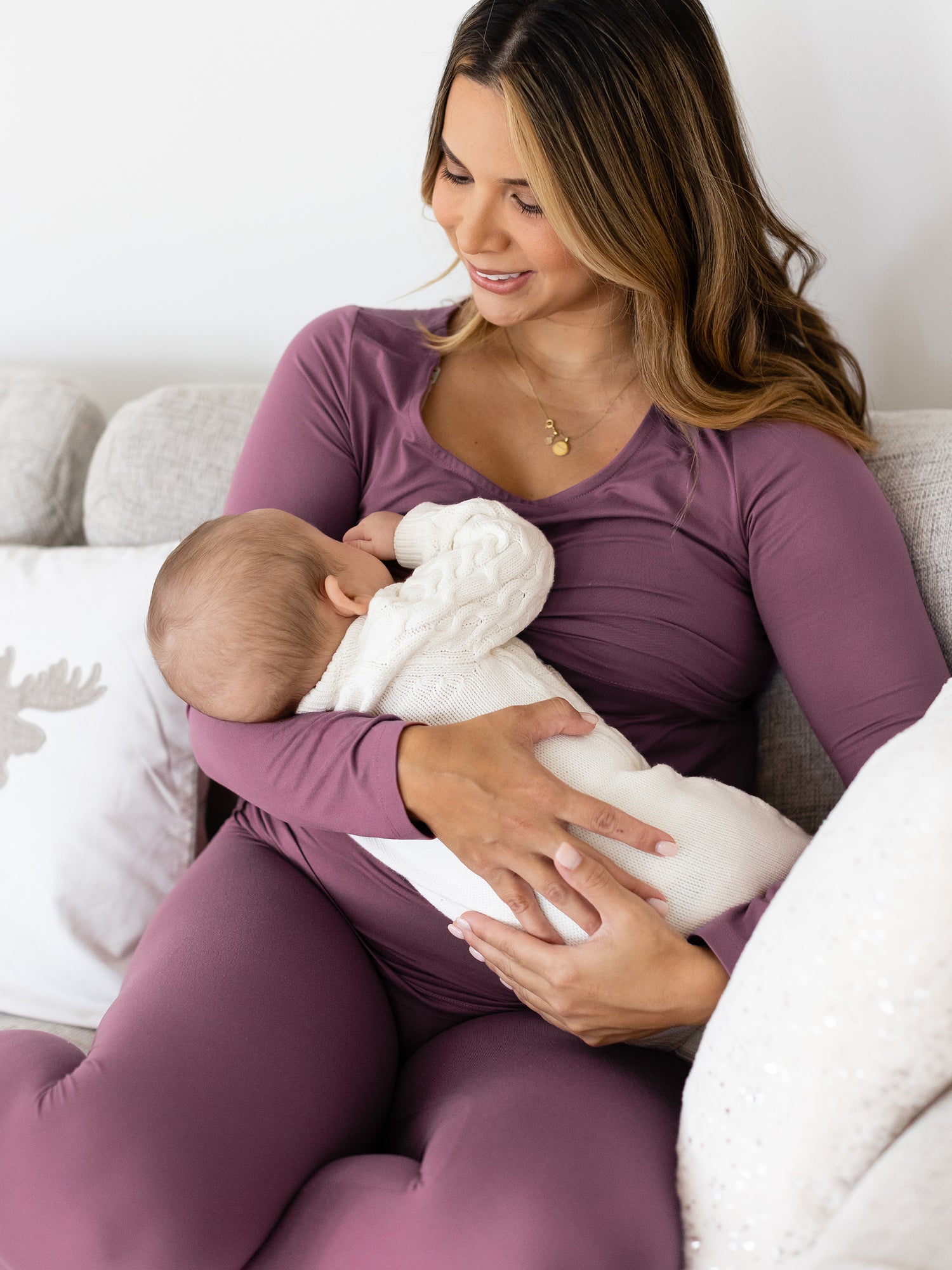 Model breastfeeding her baby while sitting on a couch wearing the jane Nursing Pajama Set in Burgundy Plum @model_info:Nicole is 5'8" and wearing a Large.