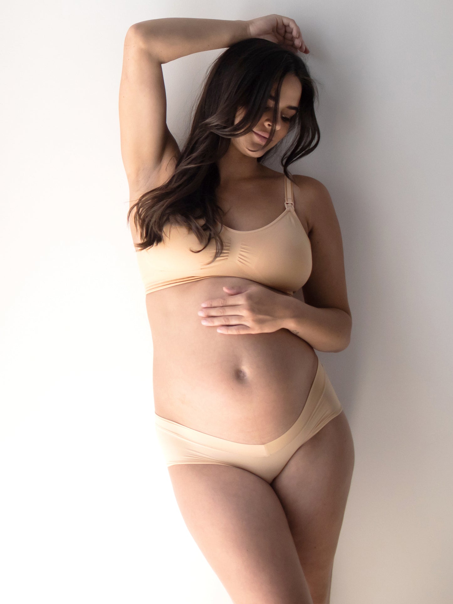 Model standing against a wall wearing the Grow with Me™ Maternity & Postpartum Hipster in Beige with her hand on her belly bump and above her head. 