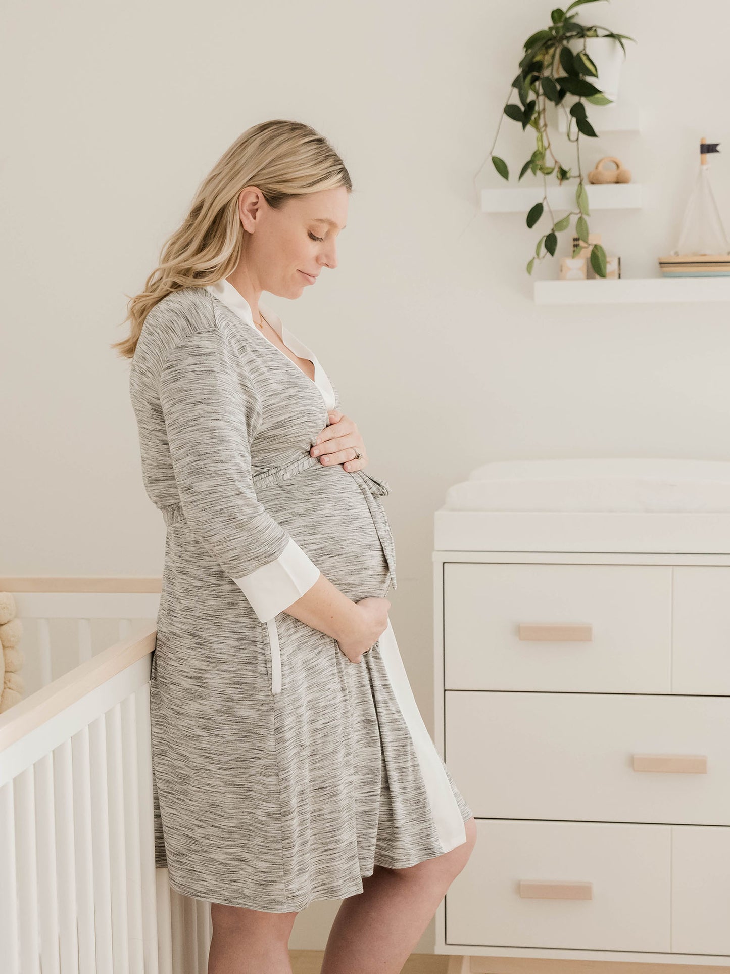 Model wearing the Emmaline Robe in Grey holding her pregnant belly in front of a crib. 