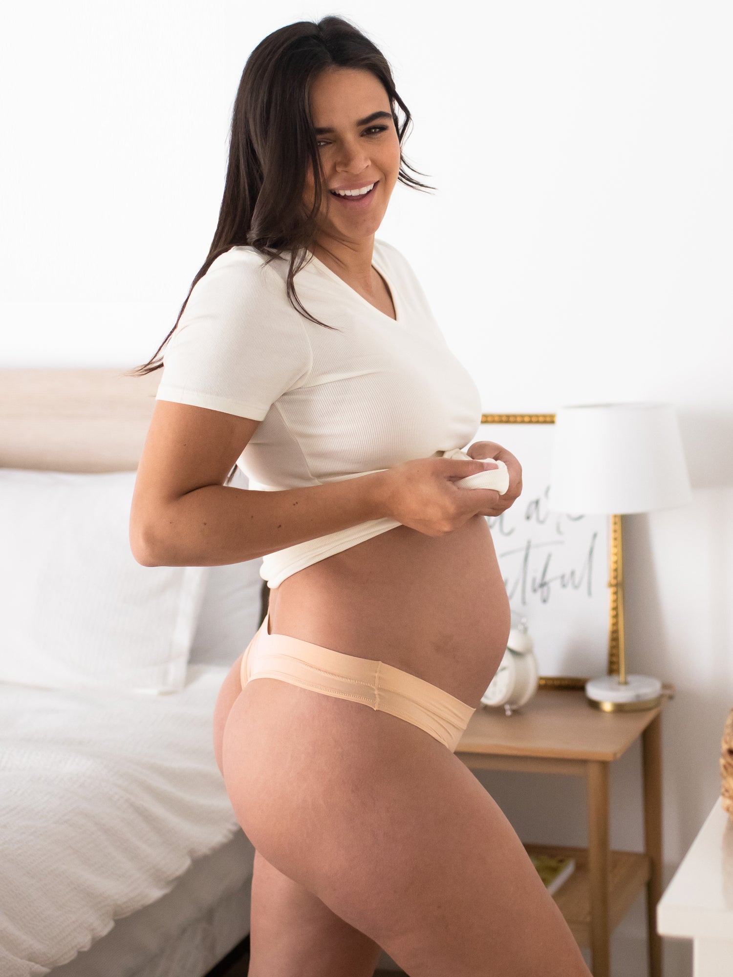 Pregnant model wearing the Grow with Me™ Maternity & Postpartum Thong in Beige holding up her shirt.