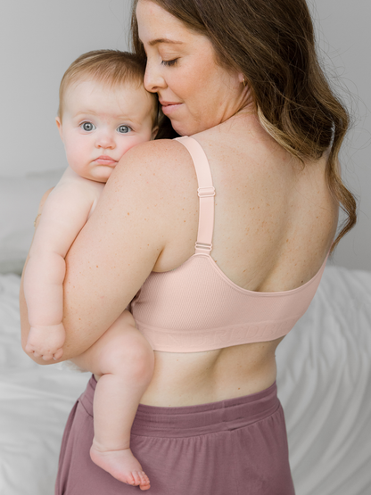 Model wearing the Sublime® Adjustable Crossover Nursing & Lounge Bra in Soft Pink holding her baby on her hip. @model_info:Ashley is wearing a Small.