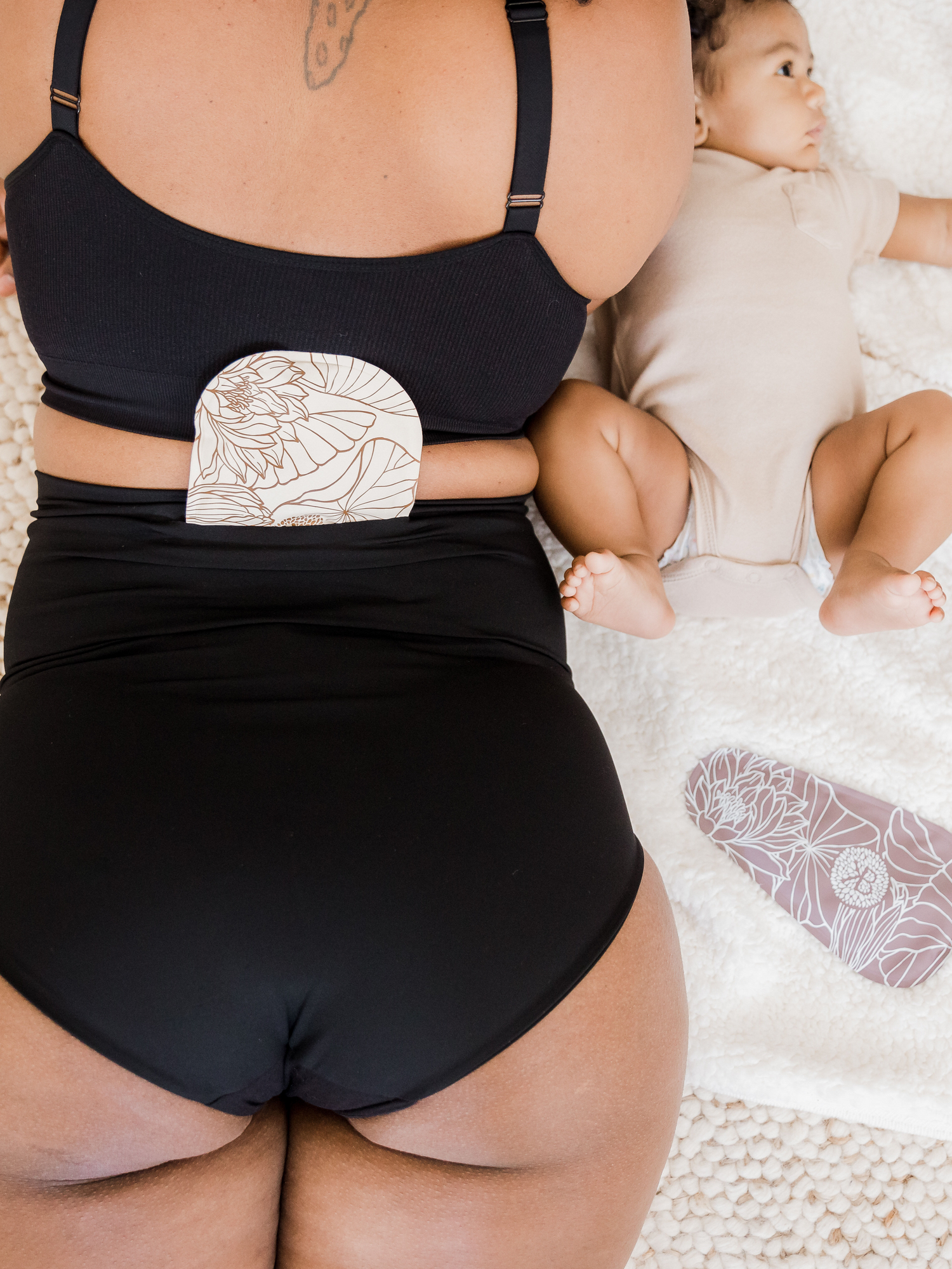 Model laying on a bed next to her baby wearing the Soothing Fourth Trimester Underwear in Black.