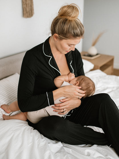 Model sitting on a bed breastfeeding her baby while wearing the Clea Bamboo Long Sleeve Pajama Set in Black.